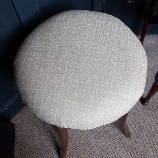 A round upholstered stool cushion in a textured cream fabric, shown from a top-down angle on a dark blue chair over a patterned carpet.